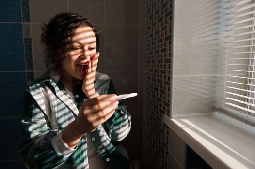 Middle aged multiracial happy woman feeling happiness while holding a positive inkjet pregnancy test, standing by the window while shadows from window blinds reflect on her face in the home bathroom