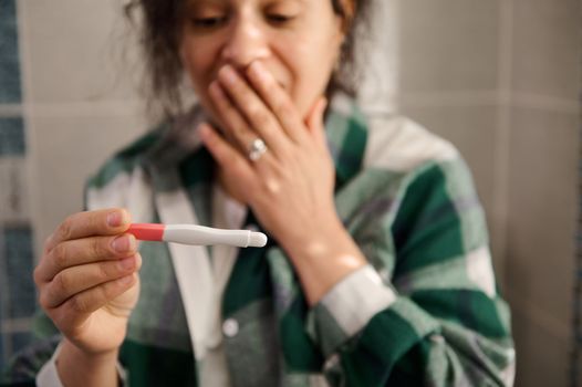 Selective focus on a pregnancy test in the hands of a blurred multi-ethnic woman, covering mouth from excitement, expressing positive emotions while rejoicing at the positive result. Finally pregnant