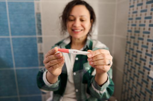 Blurred smiling middle-aged woman, expressing positive emotions and happiness, while holding out the positive pregnancy test on the camera camera. Selective focus on pregnancy test with two strips