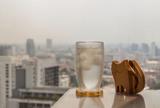 Glass of refreshing water drink with ice and wooden doll cat shape on white table in living room with city view. Place for text or advertising, Selective focus.
