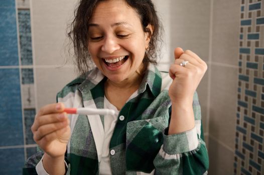 Charming woman with nervous feelings, taking pregnancy test and nervously awaiting the result, smiling from happiness while rejoicing at the positive result