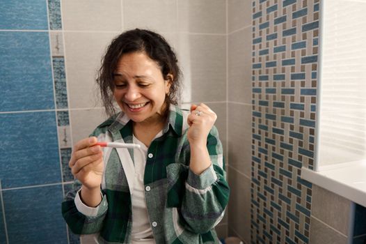 Charming multi-ethnic middle-aged woman, sitting on the toilet bowl in the home bathroom, rejoicing at positive pregnancy test. Finally pregnant. Gynecology, maternity, family and pregnancy concept