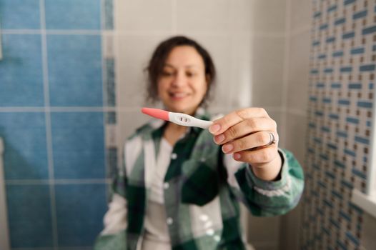 Positive pregnancy test in the hands of a blurred beautiful happy woman in the home bathroom. People, maternity and pregnancy concept