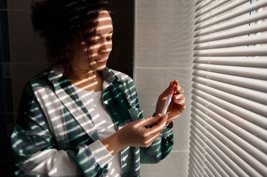 Pretty woman standing by the window and holding pregnancy test in action, awaiting the result. The shadow on of blinds on the windows falling on her, in the home bathroom. People and pregnancy concept