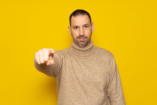 Hey, it's you. Portrait of Hispanic man wearing turtleneck sweater pointing index finger at camera, posing isolated over orange wall background. Cheerful smiling man picking, choosing and indicating