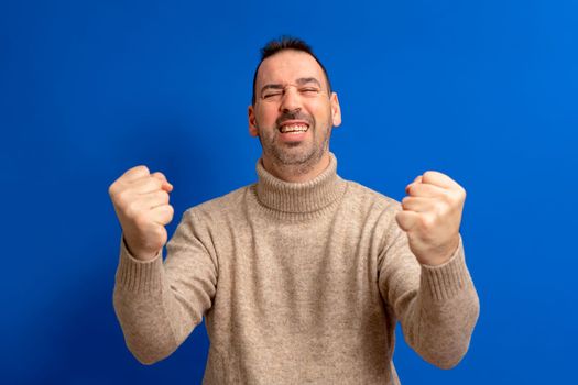Cheerful excited Hispanic man wearing turtleneck sweater standing isolated over blue background, celebrating success with fists raised and eyes closed
