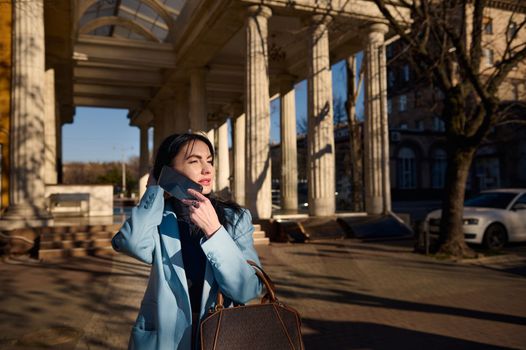 Elegant fashionable middle-aged Caucasian woman talking on mobile phone, wearing stylish blue coat, holding handbag, and the wind is blowing her beautiful long hair, while walking on spring sunny day