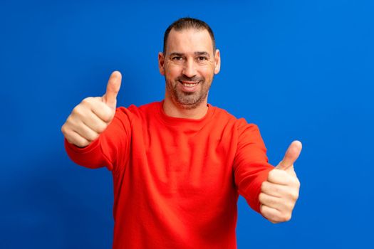 Attractive cheerful hispanic man with a beard wearing red pullover standing isolated over orange background, showing thumbs up.