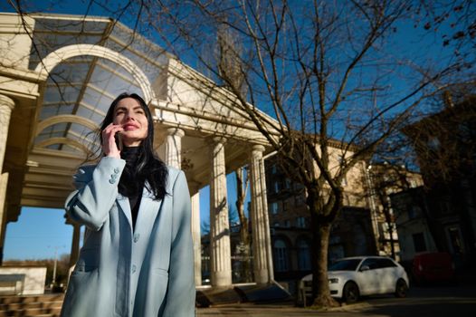 Low angle view of a beautiful Caucasian 40-44 years old woman, wearing warm blue coat, talking on mobile phone while strolling along the street on a sunny early spring day. Happy people and lifestyle