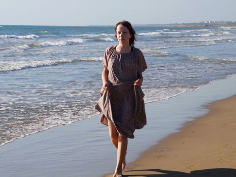 a red-haired barefoot teenage girl in a dress walks along the seashore along the water's edge.