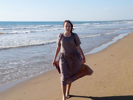 a red-haired barefoot teenage girl in a flowing dress walks along the seashore along the water's edge