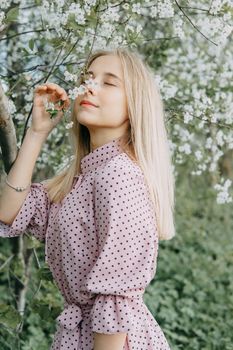 Blonde girl on a spring walk in the garden with cherry blossoms. Female portrait, close-up. A girl in a pink polka dot dress