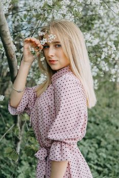 Blonde girl on a spring walk in the garden with cherry blossoms. Female portrait, close-up. A girl in a pink polka dot dress