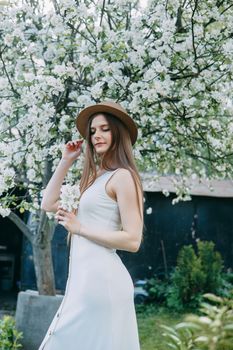 Beautiful young girl in white dress and hat in blooming Apple orchard. Blooming Apple trees with white flowers