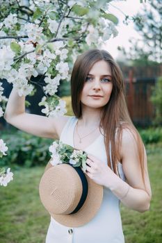 Beautiful young girl in white dress and hat in blooming Apple orchard. Blooming Apple trees with white flowers