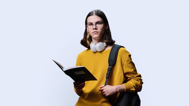 Portrait of teenage student guy on white studio background. Young male in glasses headphones holding notebook, looking at camera. High school, college, education, knowledge, science, youth concept