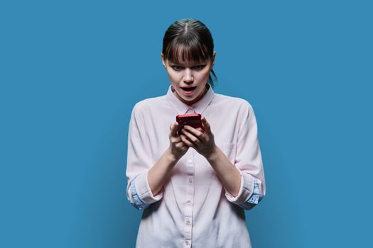 Surprised young female looking at smartphone screen on blue color studio background. Shocked excited girl reading text on phone. Emotions, lifestyle, youth concept