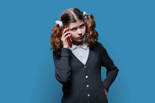 Serious preteen schoolgirl talking on mobile phone, on blue studio background. Child female student looking at camera, communication lifestyle, advertising goods and services for children