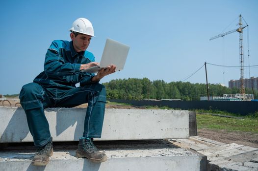 Caucasian male builder in hardhat sits on floor slabs and uses laptop at construction site