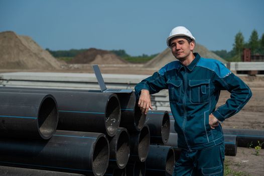 Caucasian male builder in a hard hat stands near the pipes and uses a laptop at a construction site