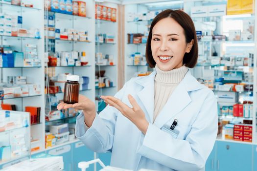 Portrait of young affable pharmacist show qualified medicine pill bottle mockup product for copyspace at pharmacy. Druggist working with her diary job at drugstore, medicine box on shelf at background