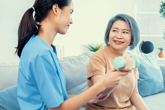 Caregiver helping contented senior woman exercise with dumbbell at home. Rehab for elder, senior medical care.