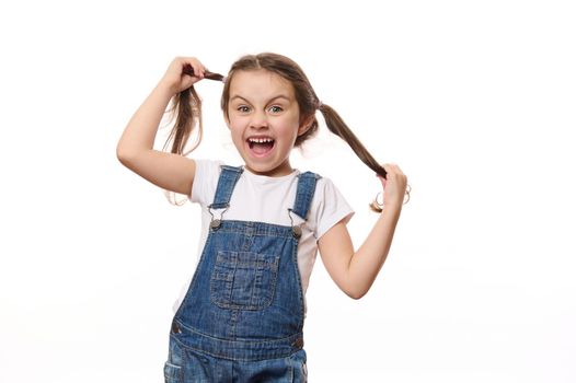 Mischievous little girl with two ponytails, wearing a t-shirt and blue denim overalls, having fun, grimacing looking at camera, isolated over white background with free space for your promotional text