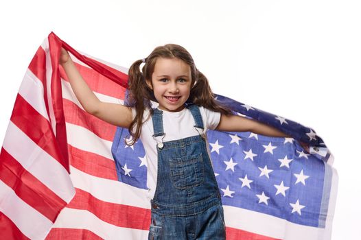 Adorable American child girl in blue denim overalls, carries USA flag, celebrates Independence Day on July 4th. Citizenship, immigration, emigration, winning the green card lottery and freedom concept