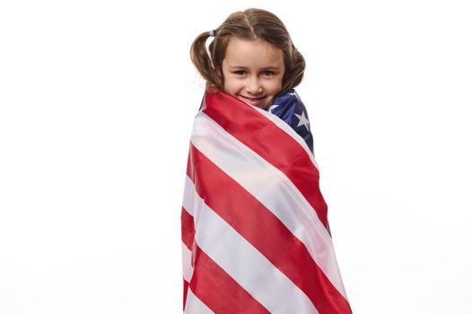 Beautiful patriotic American citizen, a lovely little child girl with two ponytails, covered in USA flag on Independance Day - 4th July, cutely smiles looking at camera, isolated over white background