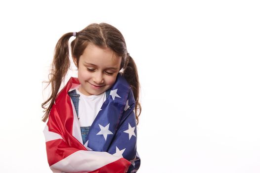 Close-up of delightful little girl with two cute ponytails, hugs herself, covered on United States of America flag, on independence day, isolated over white background, with space for your advertising