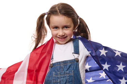Happy little American citizen, cute baby girl in blue denim overalls, with USA flag, celebrates Independence Day on July 4, isolated on white background. Immigration, citizenship and freedom concept
