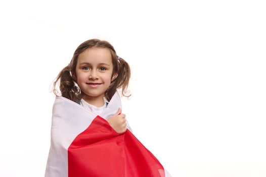 European little girl with two cute ponytails wrapping Poland flag, smiling cheerful toothy smile looking at camera, isolated on white background. Celebration of Polish Independence Day, November 11th