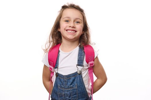 Isolated portrait on white background of a Caucasian beautiful little school child, a baby girl wearing blue denim overalls, with pink backpack, cutely smiling cheerful toothy smile looking at camera