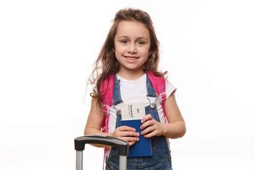 Lovely little girl in blue denim overalls, with a pink backpack, holds a boarding pass, smiles cutely looking at camera, isolated on white background with advertising space. Air travel. Trip. Tourism