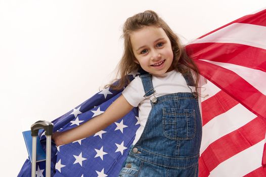 Happy baby girl wearing blue denim overalls with flag of USA, passport, boarding pass and suitcase, isolated over white background with free space for promotional text. Concept of travel, immigration