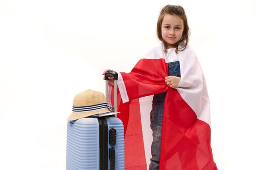 Caucasian cute little girl, little traveler, wrapping in Canada or Poland flag, smiles looking at camera, posing with blue suitcase, isolated over white background with free space for advertising text