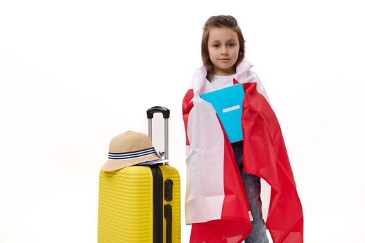Happy first grader, Caucasian little child girl wrapping in Canadian flag, holding a copybook, posing with suitcase over white background. Concept of travelling to Canada for study. Language courses