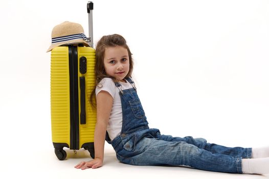 Happy little traveler girl in denim overalls, sitting leaning on her yellow suitcase, waiting to board a flight, isolated on white background. Free ad space. Journey, trip, tourism and travel concept