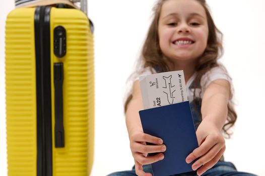 Selective focus on flight ticket or boarding pass in blurred traveler passenger child's hands near a yellow suitcase over white background. Travel. Journey. Flight. Free space for advertising text