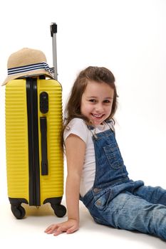 Caucasian mischievous little girl - passenger wearing blue denim overalls, with a bright yellow suitcase, smiles looking at camera, isolated over white background. Kids travelling. Journey and tourism