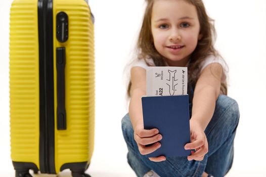 Details: boarding pass and identity documents for travel abroad in the hands of a blurred smiling little girl traveller with yellow suitcase, isolated over white background. Journey. Air flight trip