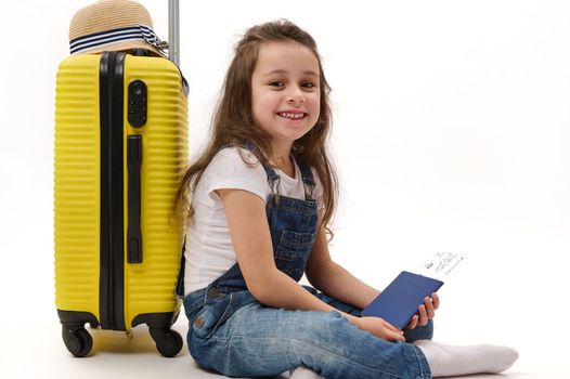 Adorable little girl passenger wearing denim overalls, sitting over white background with boarding pass in her hands, leaning on yellow suitcase and smiling a cheerful toothy smile, looking at camera