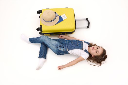 Top view cute little girl tourist in denim overalls, with suitcase, boarding pass and straw hat, lying on white background, advertising space. Travel abroad. getaway. Air flight trip journey concept