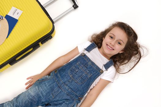 Charming European little child, adorable baby girl passenger wearing blue denim overalls, lies on white background near her boarding pass on yellow suitcase, smiles a beautiful toothy smile to camera