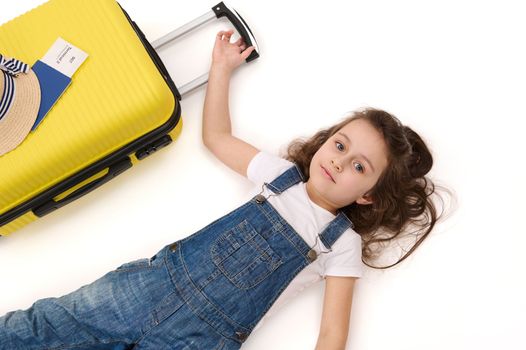 Top view of cheerful Caucasian baby girl wearing blue denim overalls, with bright yellow travel bag and boarding pass, looking at camera while waiting to board the flight, isolated on white background