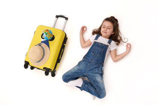 Top view of a delighted little girl in denim overalls, a traveller passenger waiting to board the flight, meditates in lotus pose, lying on white background next to her suitcase, straw hat and globe