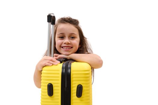 Mischievous little girl smiling a cheerful toothy smile looking at camera, leaning on yellow suitcase, isolated on white. Travel. Journey. Trip concept. Space for advertising text. Kids travelling