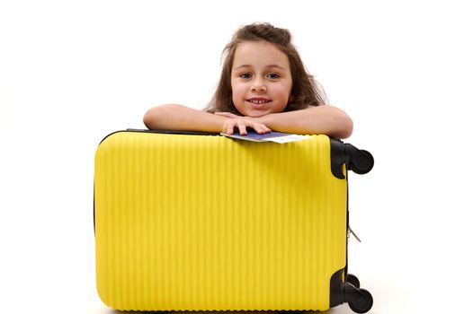 Studio shot of happy child with yellow suitcase. Portrait of cheerful kid with travel bag. Beautiful smiling Caucasian little girl with boarding pass and vacation baggage isolated on white background