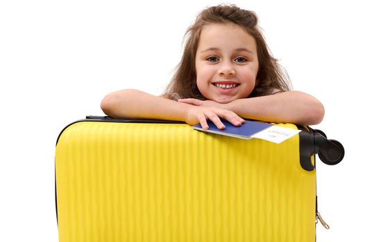 Happy little girl passenger with boarding pass, cutely smiles looking at camera, leaning on her yellow suitcase, isolated on white background. Travel. Journey. Trip. Kids travelling. Advertising space