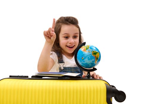 Portrait on white background of surprised little child girl with boarding pass and yellow suitcase going on summer vacation, pointing to a travel destination on globe. Holiday, travel, kids concept
